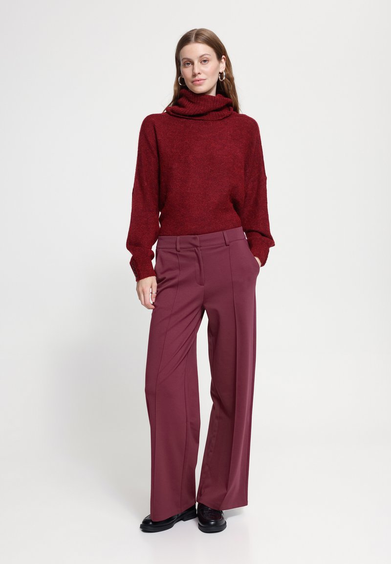 Femme portant un pull à col roulé rouge et un pantalon large mauve avec des chaussures noires, debout les mains dans les poches devant un fond uni.