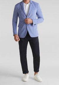 Light blue blazer with a textured finish, notched lapels, and two front pockets, paired with black pants and white sneakers.