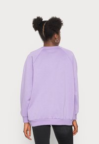 Lila mikina s raglánovými rukávy, žebrovanými manžetami a lemem. Měkká textura, jednobarevná a s minimálním zdobením vzadu.