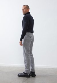 Pull à col roulé noir à côtes, pantalon gris à motif pied de poule et chaussures noires à lacets. Modèle vu de dos, se tournant légèrement sur le côté.