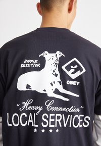 Schwarzes Baumwoll-T-Shirt mit einem weißen Hundefoto, dem Text "Hippie Detector" und "Heavy Connection Local Services" sowie Sternen darunter.
