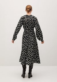 Vestido negro de mangas largas con patrón de diamantes blancos, cintura ceñida, largo midi fluido y escote fruncido. Combinado con botas negras hasta la rodilla.