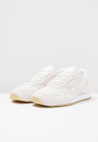 Ett par vita Reebok-sneakers med beige mockadetaljer och gummisulor på en vit yta mot en ljus, enkel bakgrund.