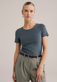 Tricou albastru cu mânecă scurtă, confecționat dintr-un material neted, cu guler rotund. Asortat cu pantaloni gri cu talie înaltă și o curea neagră.