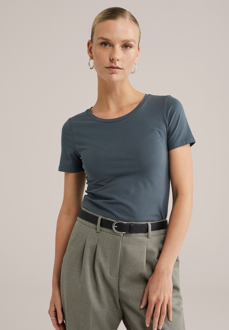 Tricou albastru cu mânecă scurtă, confecționat dintr-un material neted, cu guler rotund. Asortat cu pantaloni gri cu talie înaltă și o curea neagră.