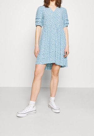Vestido azul claro con un patrón floral blanco, cuello en V, mangas cortas con volumen y cintura fruncida. Combinado con zapatillas altas blancas.