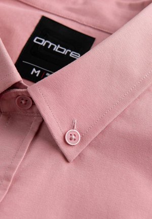 Svetle růžová košile se standartním límcem, obsahující dva odpovídající knoflíky a černou etiketu s nápisem "Ombre" bílým písmem. Hladká textura.