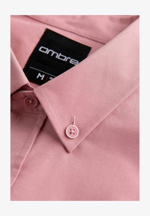 Lyserød skjorte med standard krave, prydet med to matchende knapper og en sort etikette med "Ombre" i hvid skrift. Glat tekstur.