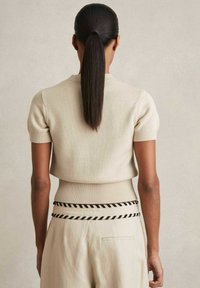 Maglione lavorato a maglia color sabbia con maniche corte e orlo a coste aderente, abbinato a pantaloni chiari caratterizzati da una cintura intrecciata bianca e nera.