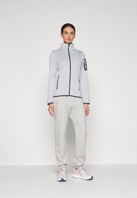 Veste grise zippée avec col montant, bordures marines contrastantes, associée à un pantalon de survêtement gris clair et des chaussures de sport avec des accents colorés.