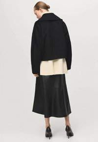 Chaqueta negra corta con cuello ancho, sobre un suéter ligero de canalé y una falda larga de cuero negra. Zapatos de tacón negros puntiagudos completan el look.