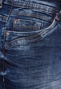 Dżinsy w ciemnoniebieskim kolorze z widocznym przeszyciem, małą przednią kieszonką i lekkim wyblaknięciem, które nadaje im zużyty wygląd. Charakteryzują się metalowym okuciem przy kieszeni.