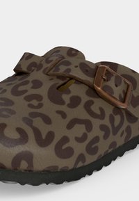 Κοντινή λήψη καφέ παπουτσιού slip-on με σκούρο λεοπάρ μοτίβο, ρυθμιζόμενο λουράκι και ορειχάλκινο αγκράφα πάνω σε μαύρη σόλα.