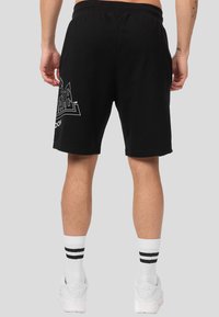 Hombre vistiendo pantalones cortos negros con un logo en el costado, zapatillas blancas Nike Air y calcetines blancos con dos rayas negras, visto desde atrás.