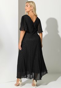 Blondynka w czarnej plisowanej sukience z przezroczystymi rękawami i złotych sandałach na wysokim obcasie, stojąca tyłem.