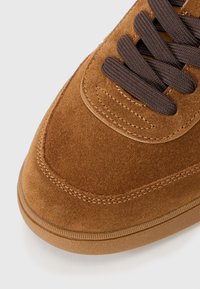 Brown suede sneaker with a textured upper, brown fabric laces, and a rubber sole featuring horizontal grooves for grip.