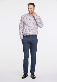 Chemise à manches longues pour homme avec un fond blanc et un imprimé floral bleu et orange. Associée à un pantalon chino bleu marine foncé et des chaussures noires.