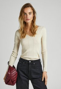 Maglione beige a coste con maniche lunghe e scollatura a V, con dettagli intricati, abbinato a jeans scuri e una borsa trapuntata bordeaux.