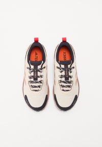 Beige sportsko med mørke detaljer, teksturert stoff, kontrasterende oransje innersåler og reflekterende detaljer, med snøring og bærehåndtak.