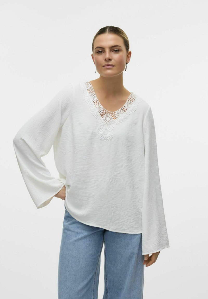 Blusa bianca a maniche lunghe con tessuto testurizzato e scollatura a V con pizzo, decorata con accenti circolari, abbinata a jeans di un blu chiaro.