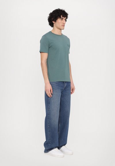 T-shirt en coton vert à manches courtes et col rond, associé à un jean en denim bleu à jambes larges et des baskets blanches. Design simple, sans motifs.