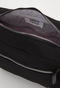 Svart Adidas-väska med silvrig dragkedja. Insidan har ett slätt, ljust foder och en etikett. Designen är snygg och minimalistisk.