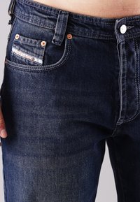 Dunkelblaue Denim-Jeans mit eng anliegendem Schnitt, ausgestattet mit kupfernen Nieten, einer kleinen Vordertasche, weißen Nähten und einem Logo-Patch über der Tasche.