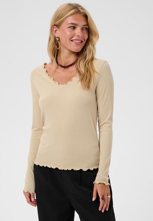 Femme blonde souriante portant un haut beige à manches longues avec des bords volantés, un pantalon noir, des boucles d'oreilles dorées, un collier de perles violet et des bagues à la main droite.