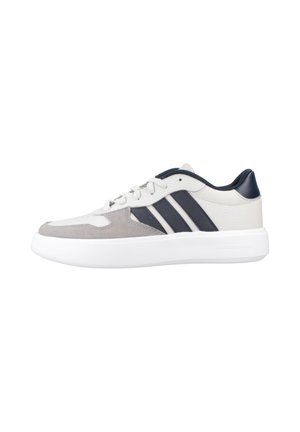 Zapatilla deportiva baja en blanco y gris con rayas negras, cierre de cordones, cuello acolchado y suela blanca gruesa.
