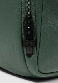 Bolso de cuero verde con cremallera negra de doble cursor, acabado texturizado y un detalle de tela visible cerca de la cremallera.