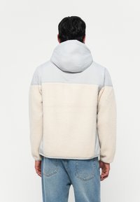 Light grey jacket with a hood, featuring a cream soft fleece panel at the back, long sleeves, and a relaxed fit.