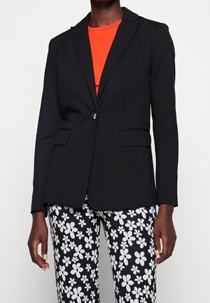 Blazer nero con rever a punta e chiusura a un bottone. Abbinato a una maglietta arancione e pantaloni neri e bianchi con motivo floreale.