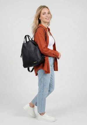 Giovane donna sorridente, indossa una camicia color ruggine, top bianco, jeans azzurri chiari, sneakers bianche, porta un grande zaino in pelle nera su una spalla.