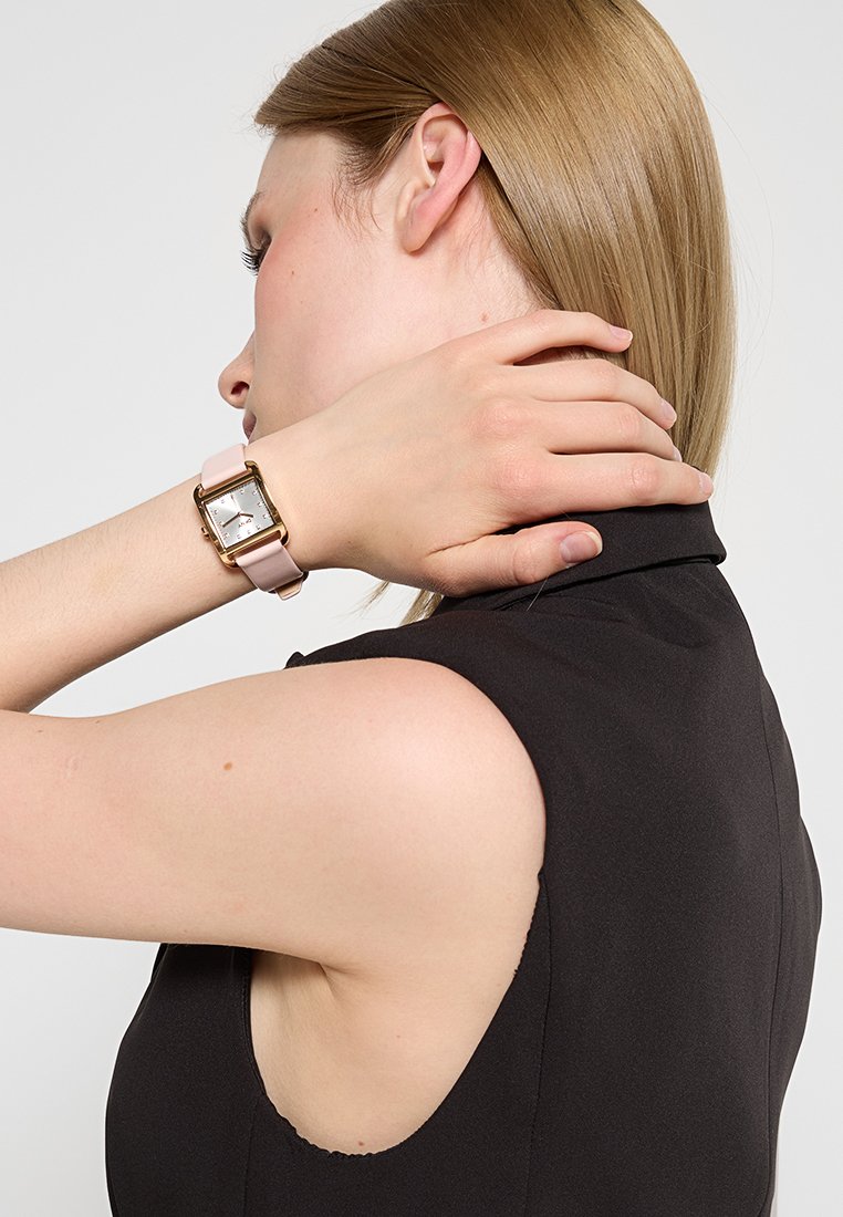 Kwadratowy zegarek z ramką w odcieniu złota, białą tarczą i różowym paskiem z silikonu. Ręka osoby poprawia włosy, ukazując szczegóły zegarka.