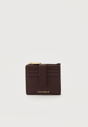 Portefeuille en cuir brun foncé avec six emplacements pour cartes, une sangle à pression, un compartiment supérieur zippé et un logo doré "Coccinelle" sur le devant.