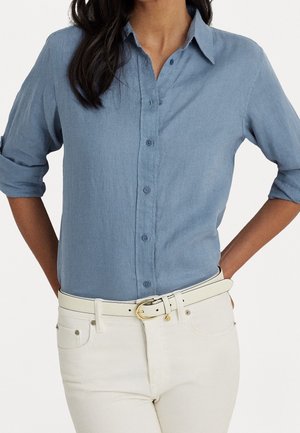 Femme portant une chemise bleue à manches retroussées, rentrée dans un pantalon blanc avec une ceinture blanche ornée d'une boucle dorée.