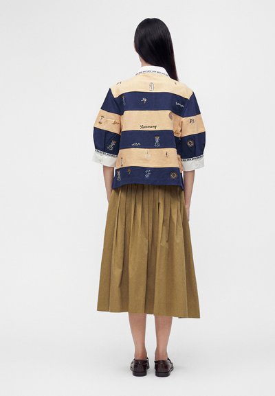 Femme aux longs cheveux foncés portant une chemise brodée rayée beige et marine, une jupe midi plissée marron et des mocassins foncés, debout, de dos.