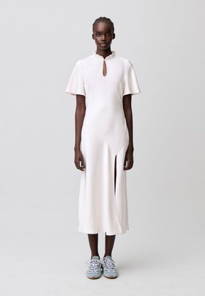 Mujer de pie con vestido midi blanco de mangas cortas, escote en ojal, abertura lateral y zapatillas deportivas azul claro sobre un fondo liso.