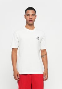 White cotton t-shirt featuring a black Adidas logo on the chest, short sleeves, and a relaxed fit. No distinct patterns or textures.