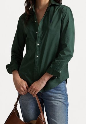 Persona con una camisa de botones verde oscuro y vaqueros azules, sosteniendo un bolso de cuero marrón con ambas manos al frente.