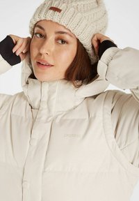 Veste rembourrée beige avec un col haut, dotée d'une fermeture à boutons et d'une texture douce. Le modèle porte un bonnet tricoté beige avec un logo en cuir.