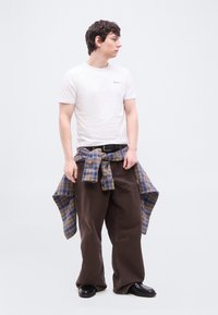 Νεαρός άνδρας που φοράει άσπρο μπλουζάκι, καφέ φαρδιά παντελόνια, μαύρα παπούτσια και ένα μπλε και καφέ καρό πουκάμισο δεμένο γύρω από τη μέση του, κοιτάζοντας προς το πλάι.