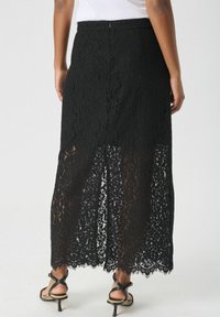 Schwarzer Spitzenrock mit transparenter Textur und gewelltem Saum, ausgestattet mit einem Schlitz hinten und einem Reißverschluss. Entworfen für eine bodenlange Passform.