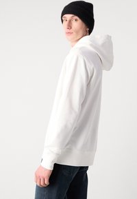Felpa bianca con cappuccio, tasca a marsupio, polsini a costine e una consistenza morbida. Abbinata a jeans in denim scuri e un berretto in maglia nera.