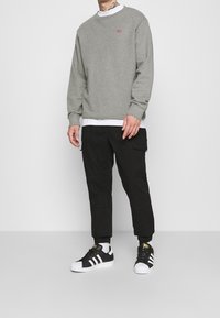 Šedá mikina s červeným logem, vrstvená přes bílou košili. Černé kalhoty typu cargo a černo-bílé tenisky. Ležérní outfit na modelce.