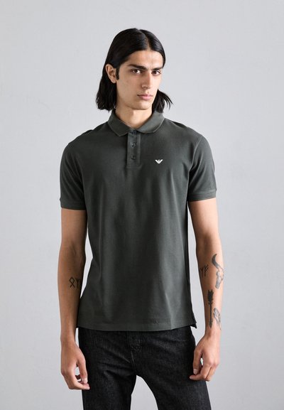 Polo shirt verde scuro in tessuto cotone, con una patta a tre bottoni, emblema del logo sul petto e maniche corte. Indossato con pantaloni in denim.