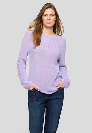 Woman with long light brown hair wearing a light purple knit sweater and dark blue jeans, standing with hands in pockets against a plain background.