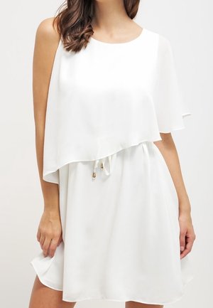 Woman wearing a sleeveless white dress with an asymmetrical layered top and a drawstring waist detail.