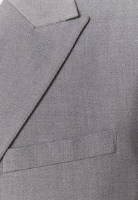 Veste de costume grise avec un tissu texturé, col cranté et poche passepoilée simple. Subtil motif de rayures verticales avec une finition nette.