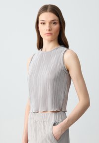 Jonge vrouw met lang bruin haar draagt een mouwloze, lichtgrijze, geplisseerde crop top en bijpassende hooggetailleerde broek, staand met één hand in de zak.